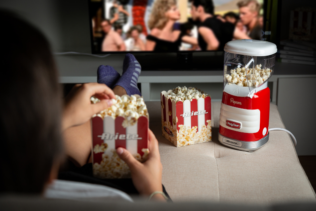 η συσκευή pop corn ακομπησμένη σε ένα μικρό τραπέζι και πίσω της φαίνεται μια ταινία στην τηλεόραση την οποία παρακολουθεί μια γυναίκα ενώ ταυτόχρονα απολαμβάνει το popcorn της.
